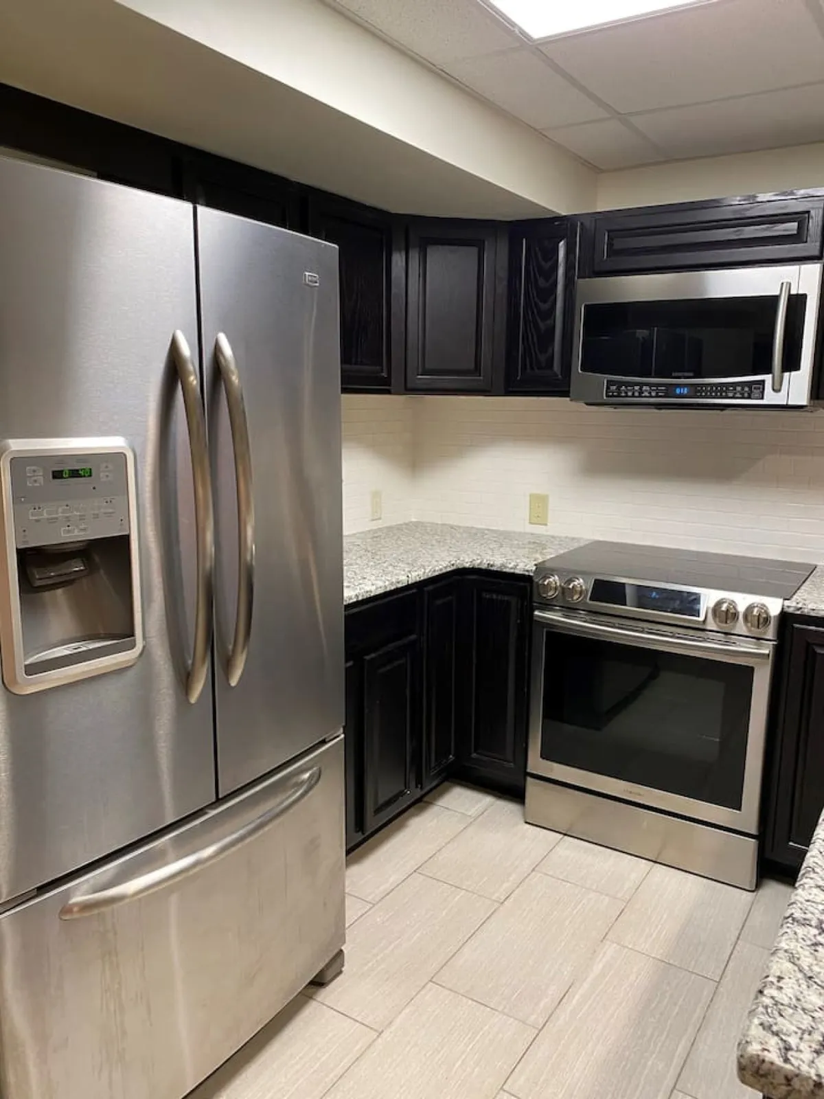 A sleek, modern kitchen area with stainless steel refrigerator and oven, granite countertops, and dark cabinetry. The contemporary ambiance invites cooking, baking, and gathering, with an open layout that encourages easy movement and interaction.