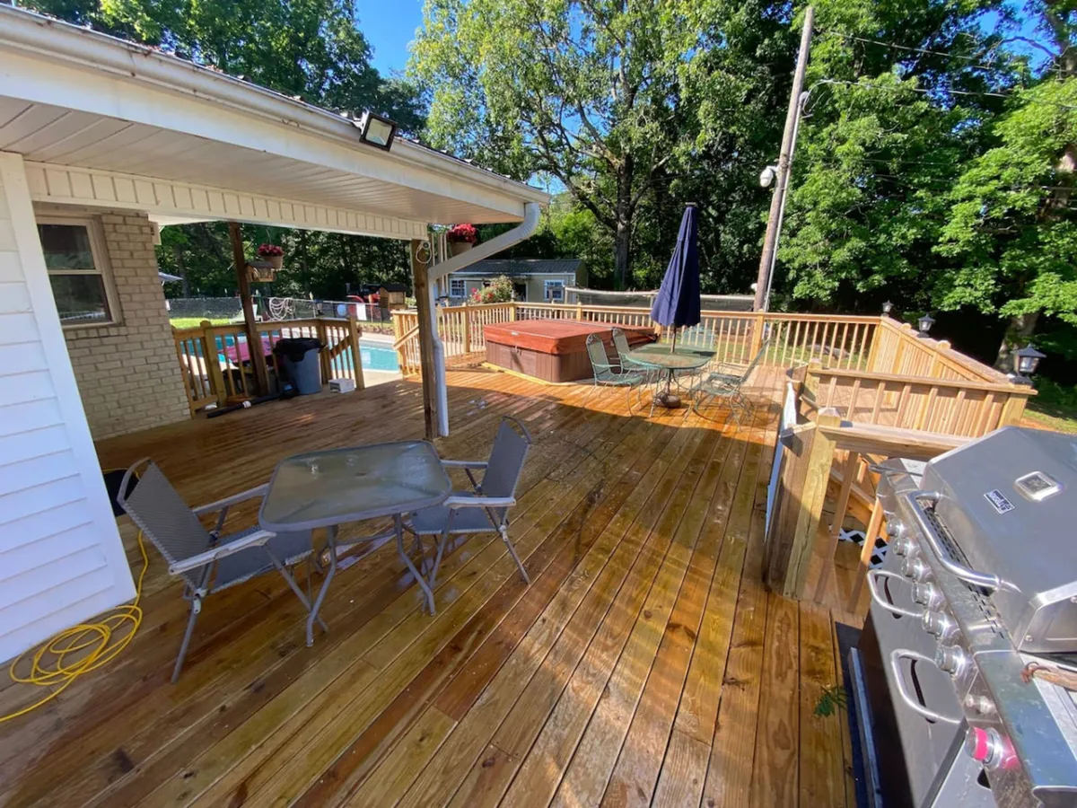 This exterior image provides a wider perspective of the pool and hot tub area, including a wooden deck, outdoor dining set, and grill. The scene is set up for social gatherings, with multiple leisure and dining options that encourage both relaxation and togetherness.