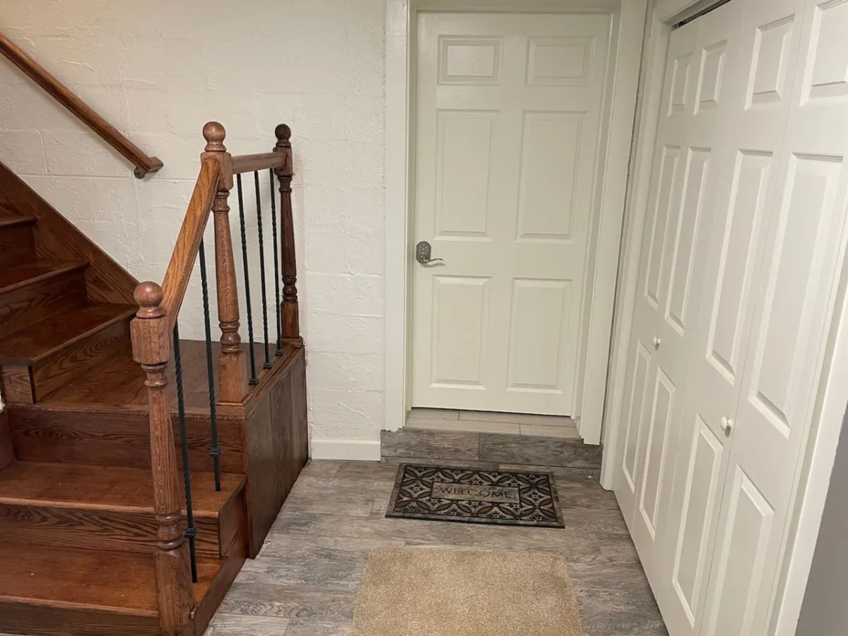 The image showcases the inviting entryway of the apartment, featuring a neatly placed welcome mat, a charming staircase, and double closet doors, all set in a warm, well-lit ambiance. The visual focus on the entry provides an immediate sense of comfort and anticipation, suggesting a homey and thoughtfully arranged interior.